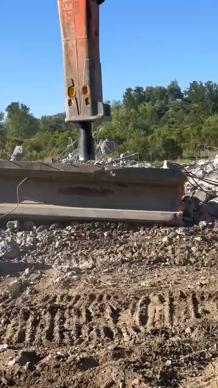 Hydraulic breaker on excavator arm hammering through a reinforced concrete slab at an outdoor demolition site with rubble and rebar visible